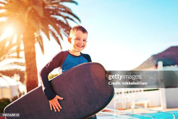 porträt von fröhlicher junge im urlaub stehen mit surfbrett neben schwimmbad - mikkelwilliam stock-fotos und bilder