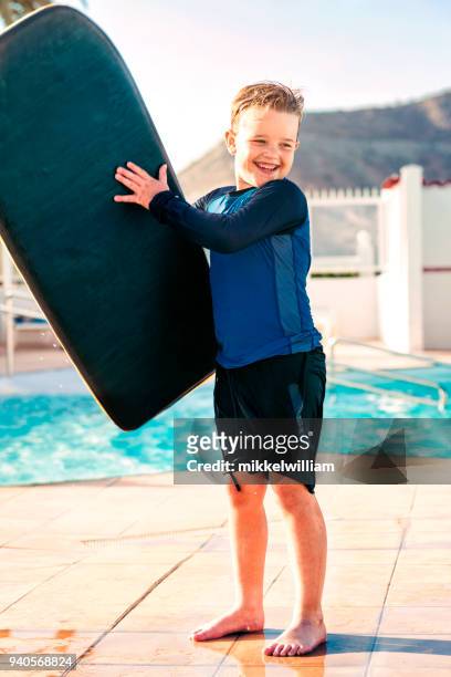 fröhlicher junge im urlaub spielt mit surfbrett neben schwimmbad - mikkelwilliam stock-fotos und bilder