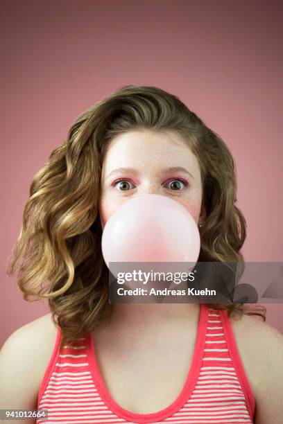 teenage girl with pale skin, freckles and wavy long hair looking into the camera with big eyes while blowing a big bubble with her pink bubblegum, portrait. - bubble gum stock pictures, royalty-free photos & images