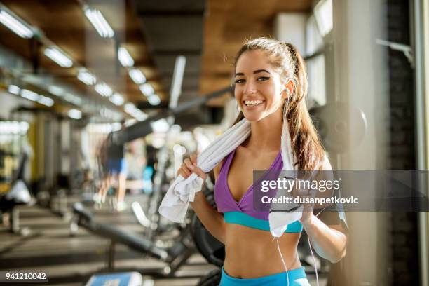 Close up portrait of adorable charming smiling young shape fitness girl with earphones holding a towel and posing while looking at the camera in the gym near the window.