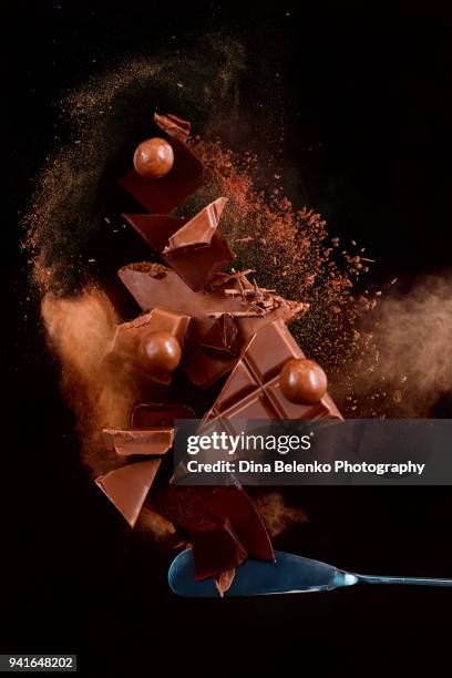 broken chocolate pieces balancing on a tip of a dessert knife with cocoa powder explosion in motion. chocolate dust on a black background. action food photography. - chocolate powder stockfoto's en -beelden