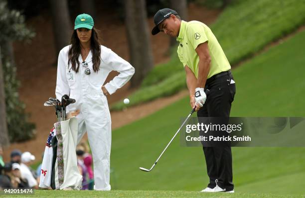 Rickie Fowler of the United States chips to a green as his girlfriend, Allison Stokke, looks on during the Par 3 Contest prior to the start of the...