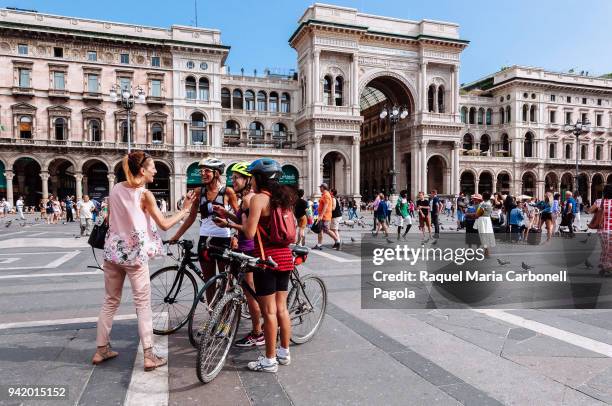 Cyclists women talking and laughing at "Piazza del Duomo" square with Galleria Vittorio Emanuele II´s triumphal arch main entrance at back.