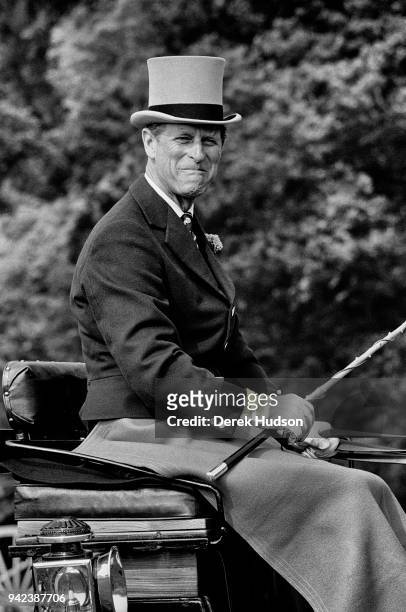 View of Prince Philip, Duke of Edinburgh, in formal attire including a top hat and gloves, as he makes a face from the driver's seat of a carriage...
