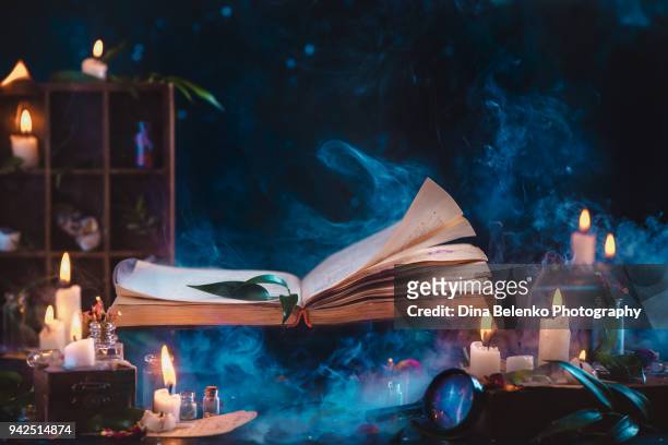 magical book of spells flying above wizard workplace with candles, herbs and a magnifying glass. dark fantasy still life with copy space. - bruja fotografías e imágenes de stock