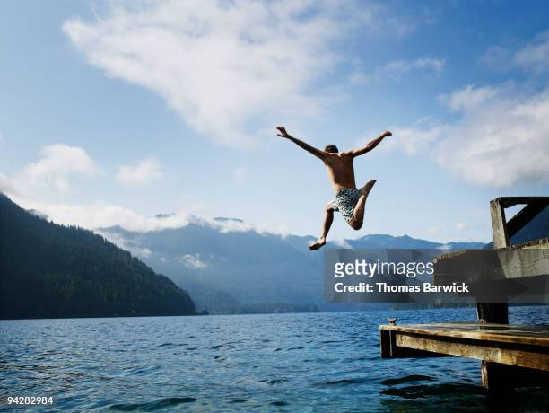 young male jumping off dock into lake in mid air - jumping into water stock pictures, royalty-free photos & images