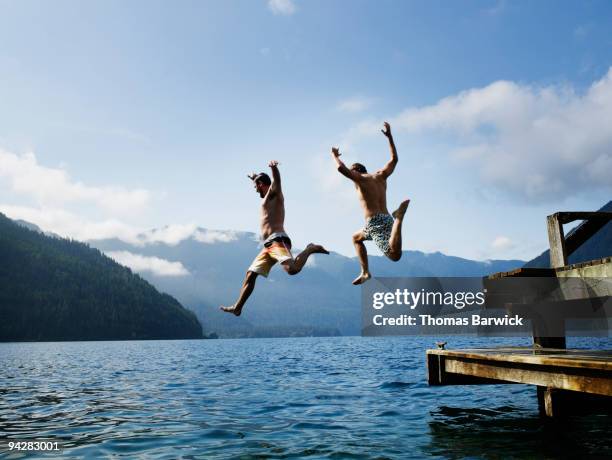 two male friends jumping off dock into lake - bergsteiger stockfoto's en -beelden