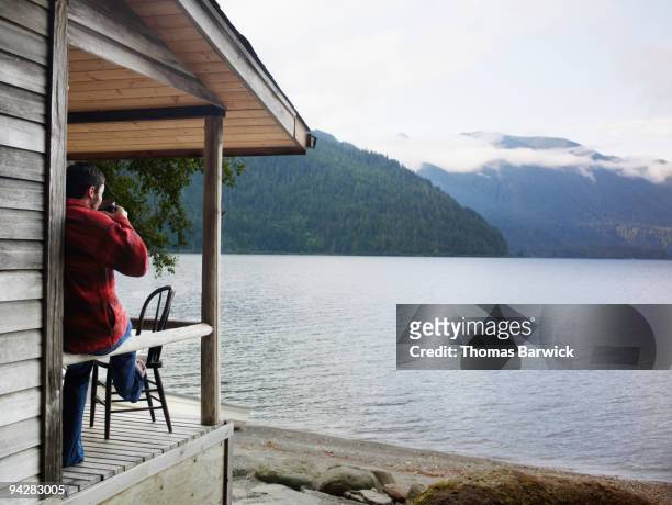 man drinking coffee on front porch of lake cabin - cabaña de madera fotografías e imágenes de stock