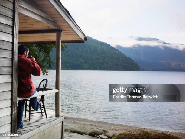 man drinking coffee on front porch of lake cabin - log cabin stock pictures, royalty-free photos & images