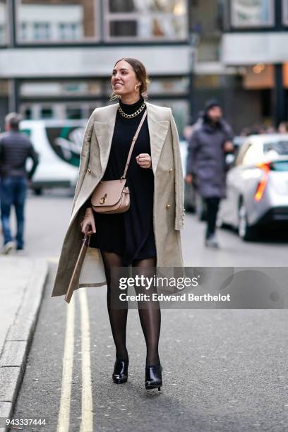 Guest wears earrings, a beige coat, a pink bag, a black dress, a golden necklace, black tights, black shoes, during London Fashion Week February 2018...