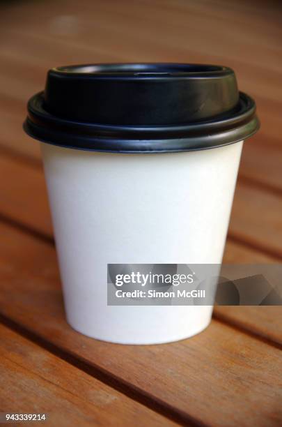 a disposable white cardboard coffee cup with a black plastic lid in sunlight on a wooden picnic table - coffee cup takeaway stock pictures, royalty-free photos & images