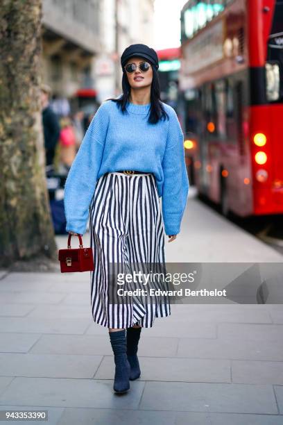 Guest wears a hat, sunglasses, blue oversized pull over, a black and white striped skirt, a red bag, shoes, during London Fashion Week February 2018...