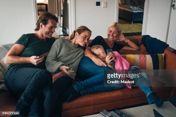 smiling family looking at boy using mobile phone while sitting on sofa at home - children phone couch stock pictures, royalty-free photos & images