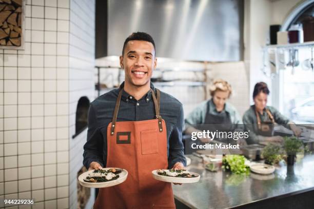 portrait of smiling waiter holding fresh food plates against female chefs in restaurant kitchen - servitör bildbanksfoton och bilder