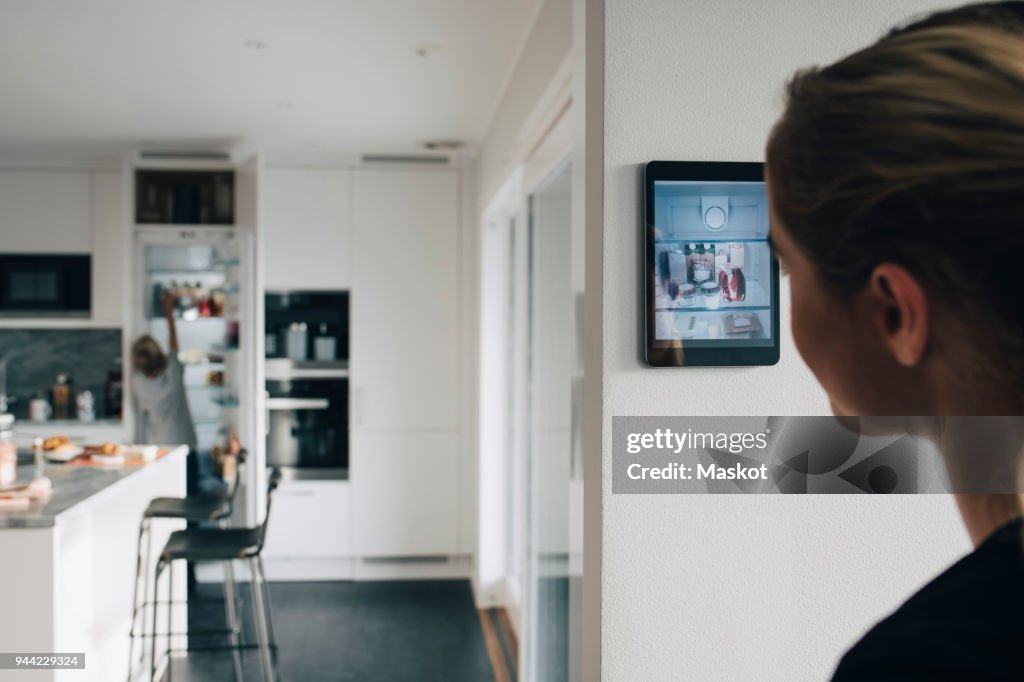 Rear view of teenage girl standing by wall with digital tablet looking at brother in kitchen