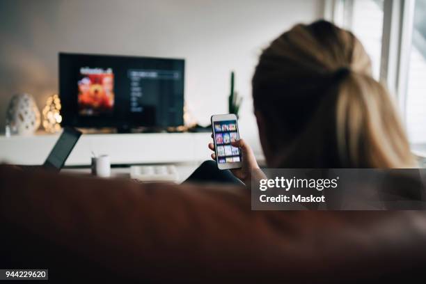 rear view of teenage girl using smart phone app while watching tv in living room at home - watch stockfoto's en -beelden