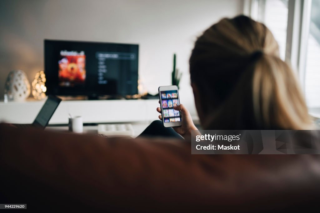 Rear view of teenage girl using smart phone app while watching TV in living room at home