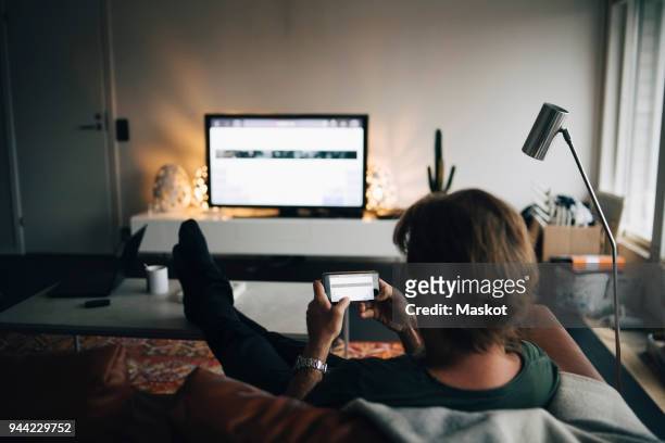 full length of man using mobile phone while sitting on sofa in living room - man watching tv alone stock-fotos und bilder