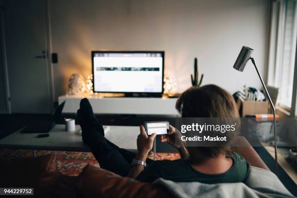 full length of man using mobile phone while sitting on sofa in living room - guy with phone full image ストックフォトと画像