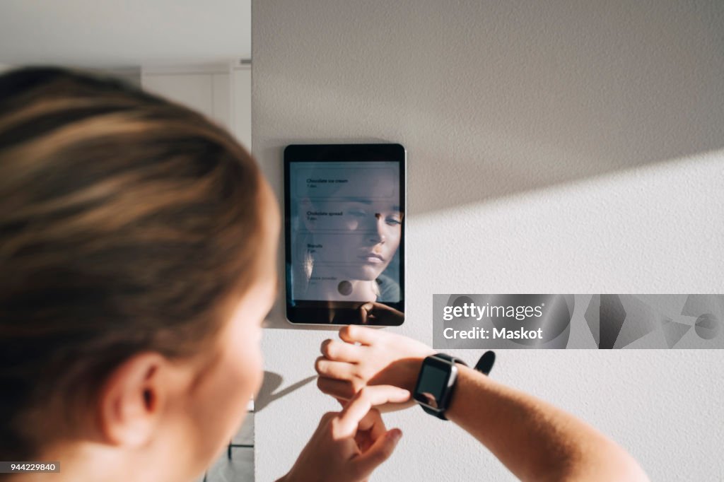 Teenage girl adjusting smart watch with digital tablet mounted on white wall at home