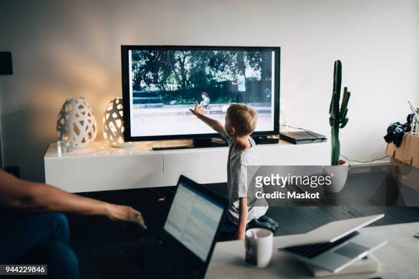 full length of boy kneeling while touching smart tv in living room at home - familia viendo tv fotografías e imágenes de stock