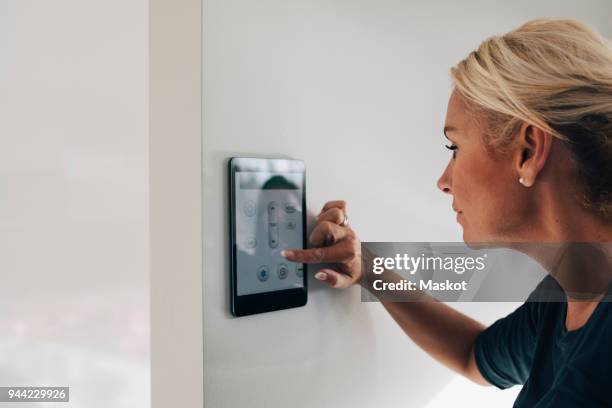 blond woman adjusting thermostat using digital tablet mounted on white wall at home - thermostaat stockfoto's en -beelden
