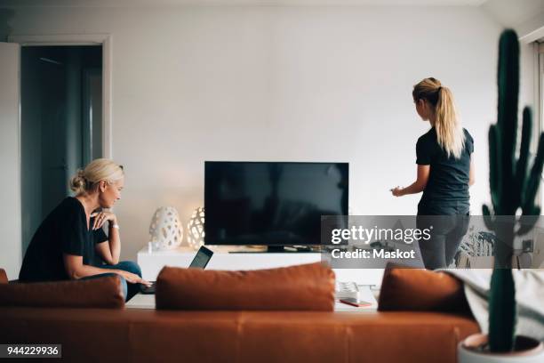 woman using laptop while teenage daughter standing by television set in living room at home - living room people no tv stock pictures, royalty-free photos & images