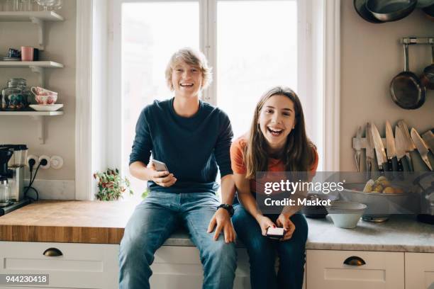 portrait of male and female friends with mobile phones sitting at kitchen counter - boy kitchen stockfoto's en -beelden