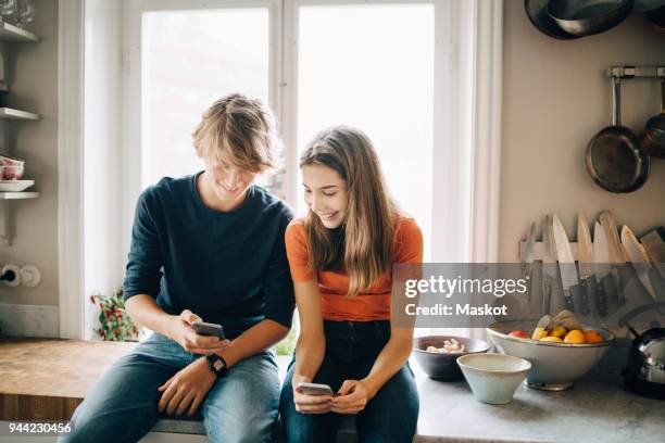 teenage boy showing mobile phone to female friend while sitting at kitchen counter - kid presenting stockfoto's en -beelden