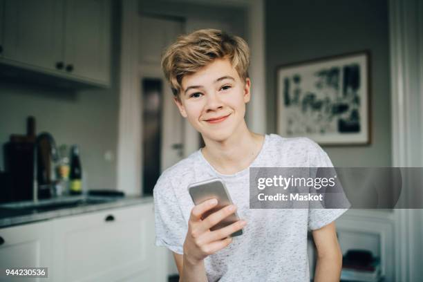 portrait of smiling teenage boy holding smart phone while sitting in kitchen at home - teenage boys stock pictures, royalty-free photos & images