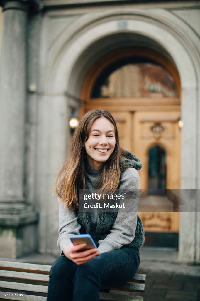 Portrait of smiling girl with smart phone sitting on bench in city
