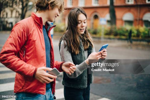 girl talking while showing mobile phone to male friend on city street - boy and girl talking fotografías e imágenes de stock