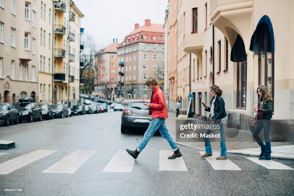 Full length of friends using mobile phone while crossing street in city