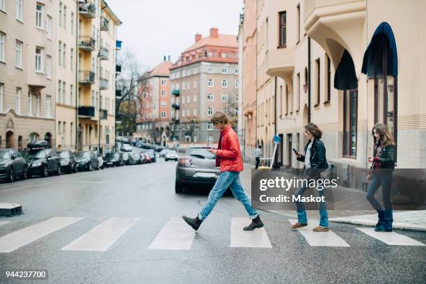 full length of friends using mobile phone while crossing street in city - crossing imagens e fotografias de stock