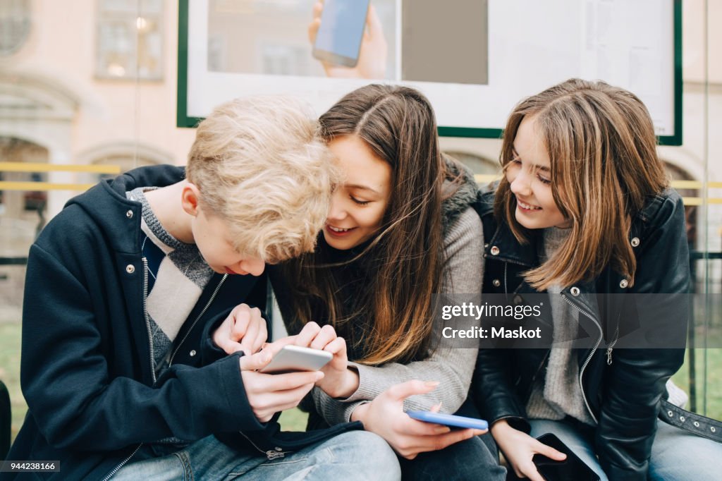 Smiling friends using mobile phone while sitting at bus stop