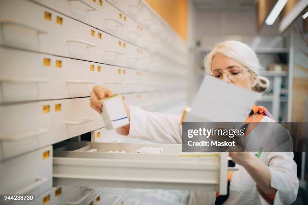 mature female pharmacist holding prescription paper checking medicine by drawer in store - apothekerin stock-fotos und bilder