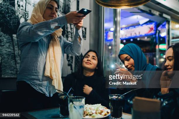 Young Muslim females photographing food and drinks on table with friends at cafe