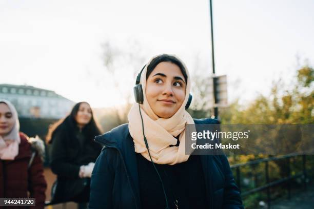 smiling teenage girl listening to headphones with friends against sky - listening stockfoto's en -beelden