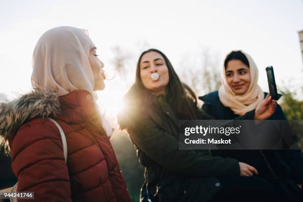 multi-ethnic female friends blowing bubble gums while taking selfie against sky - 3 teenagers mobile outdoors stock-fotos und bilder