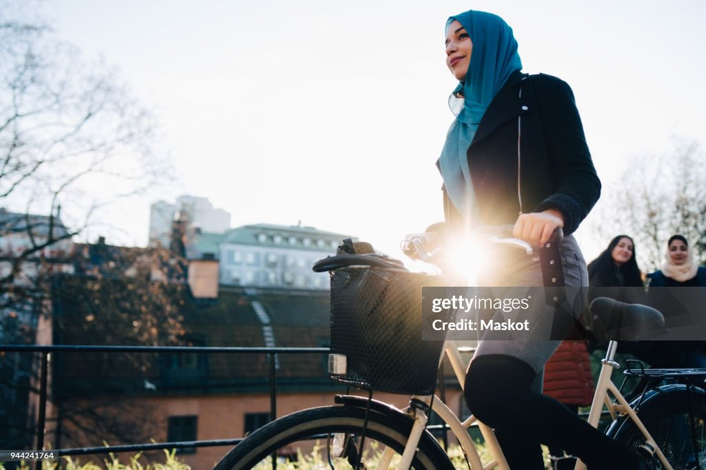 Back lit of young woman cycling by female friends against sky in city