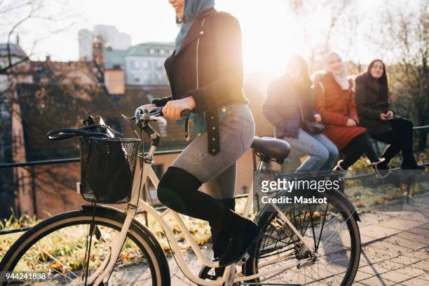 low section of woman cycling by female friends sitting on railing in city - low section stockfoto's en -beelden