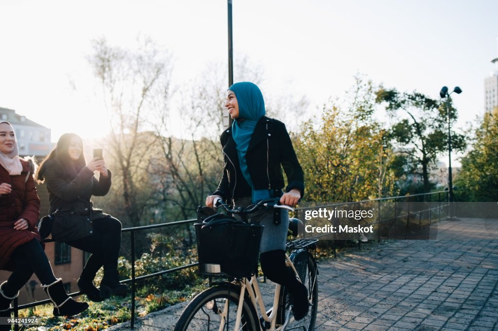 Smiling young woman cycling on footpath by female friends sitting on railing in city