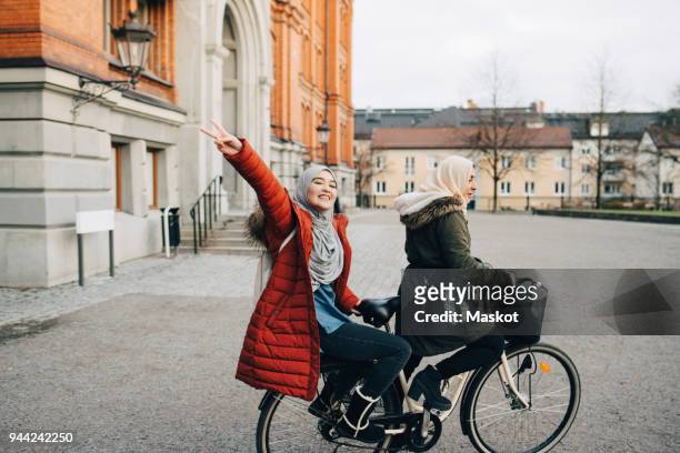 portrait of happy young woman showing peace sign sitting behind friend on bicycle in city - holy city stock pictures, royalty-free photos & images