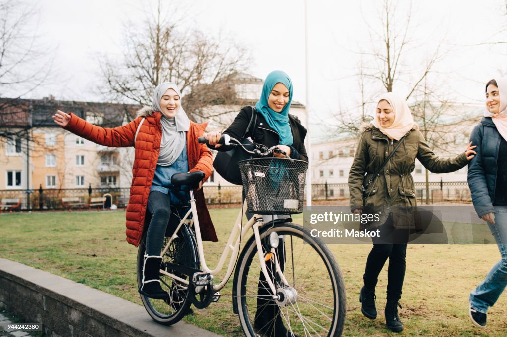 Cheerful multi-ethnic female friends walking with bicycle on grass in city