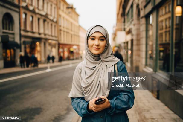 portrait of confident young woman wearing hijab standing with mobile phone on sidewalk in city - velo fotografías e imágenes de stock