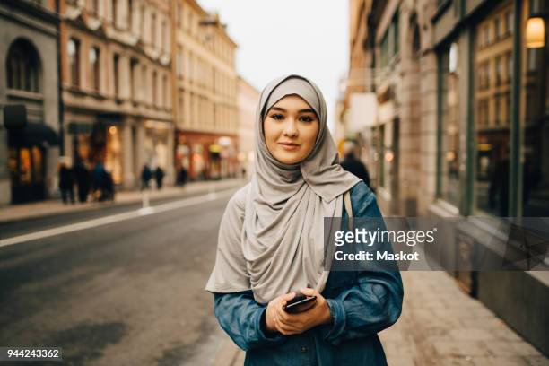 Portrait of confident young woman wearing hijab standing with mobile phone on sidewalk in city