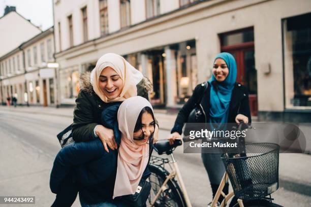 cheerful teenage girl giving young woman piggyback by friend walking with bicycle on street in city - young muslim stockfoto's en -beelden