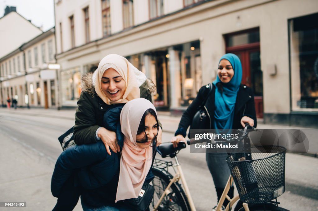 Cheerful teenage girl giving young woman piggyback by friend walking with bicycle on street in city
