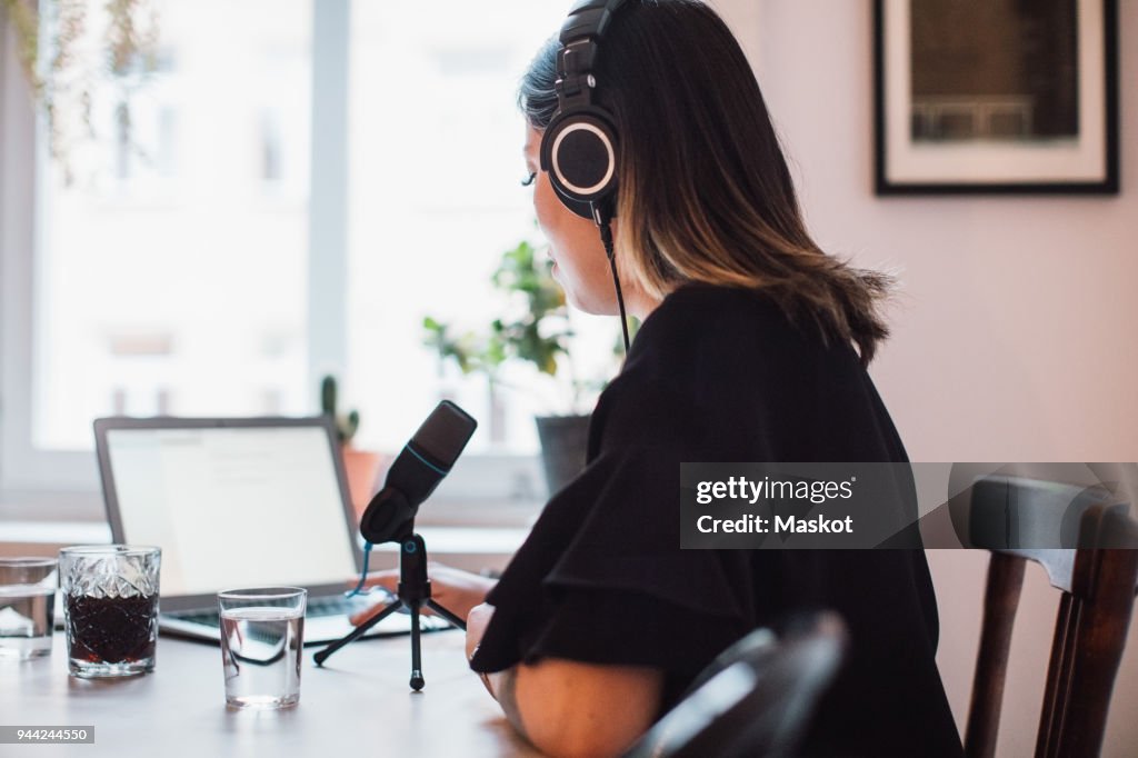 Side view of female podcaster wearing headphones sitting at table with microphone at home