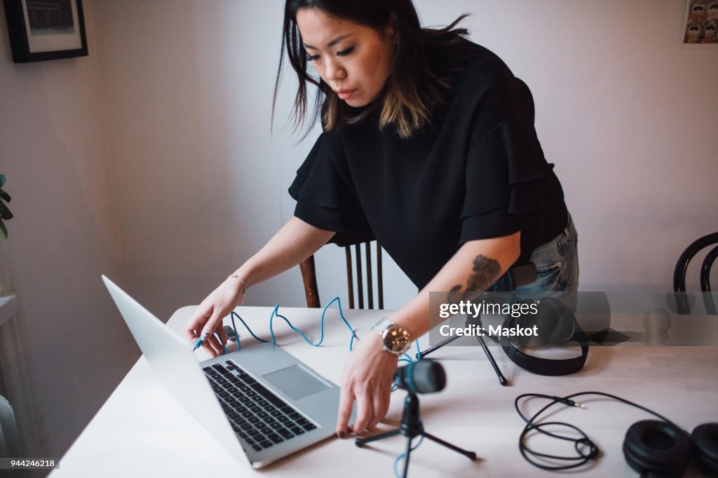 Female blogger using laptop standing by table with microphone at home